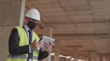 Düşük açılı beyaz erkek inşaat yöneticisi yüz maskesi takıyor, yeşil yansıtıcı yelek ve beyaz başlık takıyor, tablet bilgisayar tutuyor, sonra da kameraya bakıyor.
