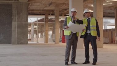 İki beyaz erkek inşaat işçisinin resmi giysiler, yeşil yansıtıcı yelekler ve beyaz başlıklar giyip binada dikilip, projeyi tartışırken geniş açıdan çekilmiş görüntüleri.