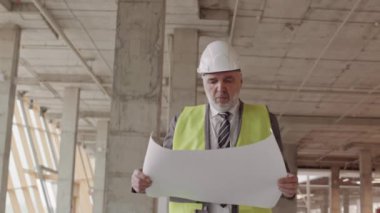 Beyaz saçlı, resmi takım elbiseli, yansıtıcı yelek ve beyaz şapka giyen beyaz saçlı bir ustabaşı, elinde proje planı olan büyük bir kağıt tutarak binanın içinde yürüyor.