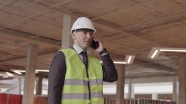 Resmi takım elbise giyen, yansıtıcı yelek ve beyaz şapka takan, cep telefonuyla konuşan, binanın içinde yürüyen Kafkas ustabaşısının izini sürme aracı.