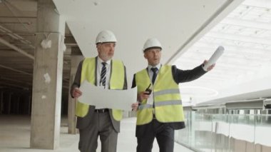 Üzerinde yansıtıcı yelek ve beyaz başlıklar olan resmi kıyafetler giyen iki Kafkas işadamının orta uzunluktaki takibi, proje planlı kağıt tutmaları, alışveriş merkezinin yapımını denetlemeleri, konuşmaları...