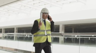 Beyaz bir mimarın resmi bir takım elbise, yansıtıcı yelek, koruyucu gözlük ve miğfer takması, elinde kağıt tutması, akıllı telefonla konuşması, şantiyede dolaşması.