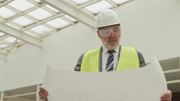 低角的白种人工头 身穿绿色反光背心 头戴硬礼帽 站在建筑工地上 仔细观察造纸工程 — 图库视频影像