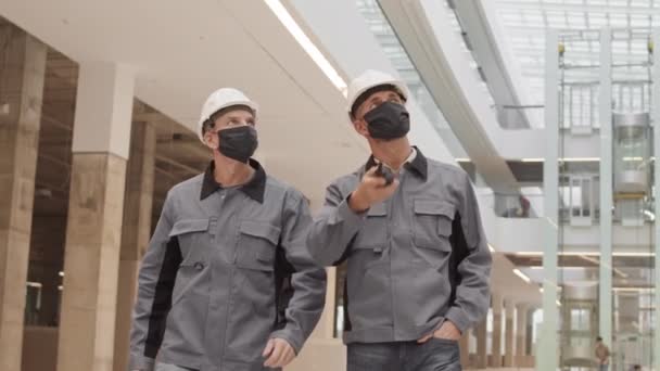 白い硬い帽子と顔のマスクを身に着けている2人の白人男性の中程度の追跡 建設中の大規模な建物の中を歩く 周りを見回す — ストック動画