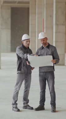 Kafkasyalı ustabaşının inşaatçılara proje planını açıklarken tam dikey çekimi, inşaat alanında duran işçi ceketi ve başlığı giyen adamlar.