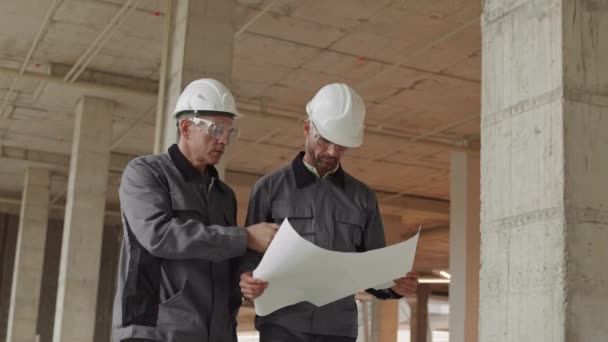 两名白人男子身穿工人夹克 头戴安全帽 戴着安全帽 在建筑工地走来走去 一名男子持纸 他的同事 对讲机 — 图库视频影像