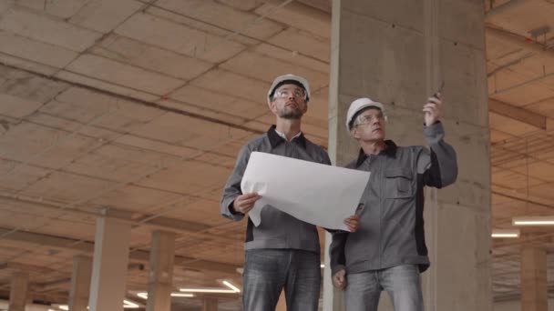 两名白人工人 身穿工作服 头戴安全帽 头戴安全帽 站在建筑工地 手里拿着一张纸 手里拿着他的同事 对讲机 — 图库视频影像