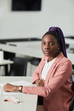 Sınıfta masa başında otururken okulda kameraya bakan Afro-Amerikan genç bir kadının minimum dikey portresi