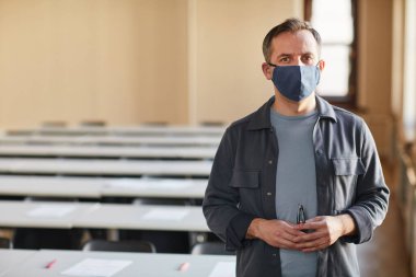 Oditoryumda dikilip kameraya bakarken maske takan olgun üniversite profesörünün belden yukarı portresi.