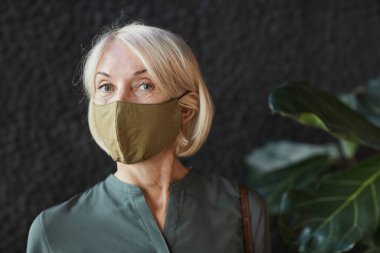 Çekici sarı saçlı, bez maskeli, koyu renk bluzlu, siyah arka planda bitki örtüsü olan son sınıf beyaz kadın portresi.