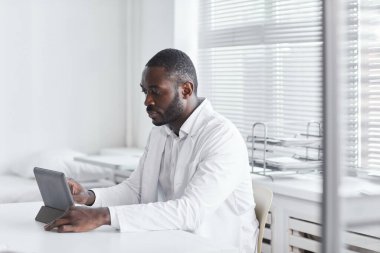 Afrikalı genç doktor masada oturuyor ve ofisinde dijital tablet üzerinde çalışıyor.