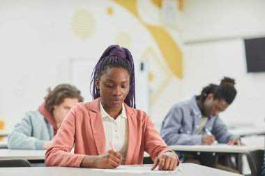 Okul sınıfında çalışan çeşitli öğrenci grupları ön masada oturan Afro-Amerikan genç bayana odaklanıyor.