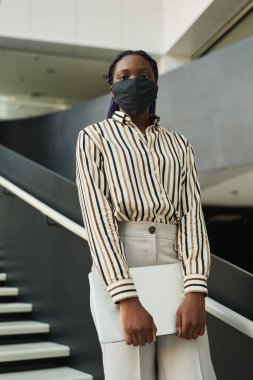 Ofiste poz verirken maske takan ve kameraya bakan kendine güvenen Afro-Amerikan iş kadınının dikey portresi