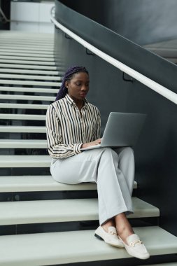 Ofiste merdivenlerde otururken dizüstü bilgisayar kullanarak siyah beyaz giyinmiş Afro-Amerikalı bir iş kadınının tam boy portresi.