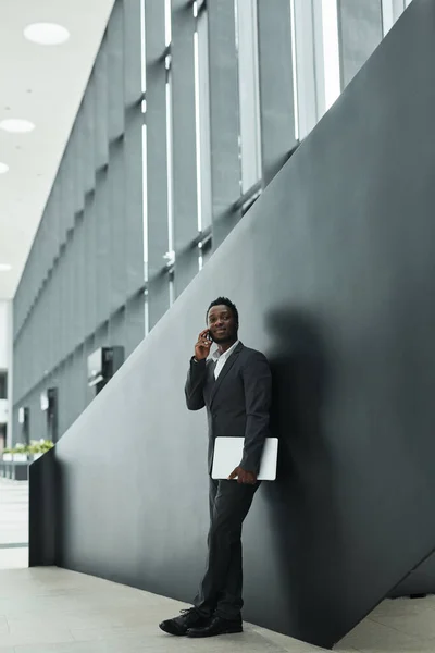 Graphic wide angle portrait of confident African-American businessman speaking by smartphone while standing against black background in office hall