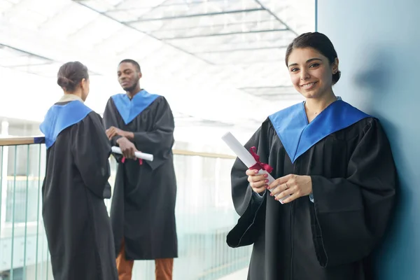 卒業生を身に着けている若者のグループは 現代の大学のインテリアで屋内でガウンを着て 笑顔に焦点を当て中東の女性は卒業証書を保持し 前景にカメラを見て スペースをコピー — ストック写真