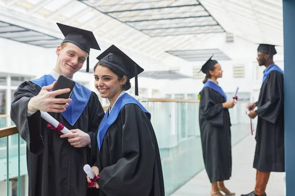 2人の幸せな若者が卒業生のガウンを着用し 大学のインテリアで屋内で自撮りしながらスマートフォンを保持 — ストック写真