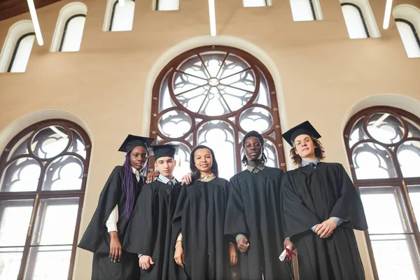 古典的な学校の講堂で華やかな窓のそばに立っている間 卒業衣を着た若者の多様なグループでの広い角度ビュー コピースペース — ストック写真