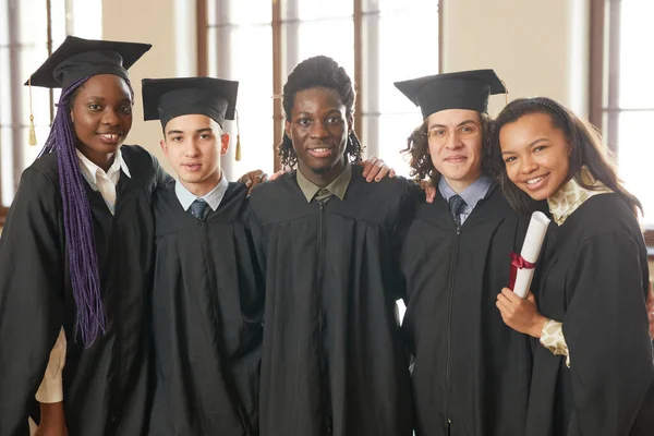 学校の講堂に立ちながら卒業衣を着てカメラで微笑む若者たちの多民族集団の腰像 — ストック写真