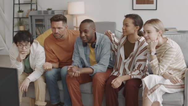 自宅のリビングルームでソファに座っている5人の若い多民族の友人の中長期 テレビを見て 密かに白人男性の肩にタッピング混合レースの女性 — ストック動画