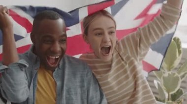 Orta boy, neşeli, genç Afrikalı erkek ve beyaz bir kadının İngiliz bayrağını sırtında taşıdığı yakın plan çekimler. Sonra da evdeki favori spor takımı için tezahürat yapıyorlar.