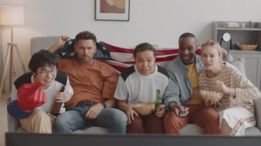 Orta uzunlukta etnik çeşitlilikte kadın ve erkek arkadaşlar evlerindeki kanepede oturup, bira ve atıştırmalıklar yiyip, favori takımın kazanmasına rağmen Amerikan bayrağını sallayıp alkışlıyorlar.