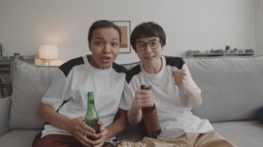Orta boylu Asyalı bir adam ve aynı tişörtleri giyen, oturma odasındaki kanepede oturan, favori takımın zaferini bira eşliğinde kutlayan melez bir kadın.