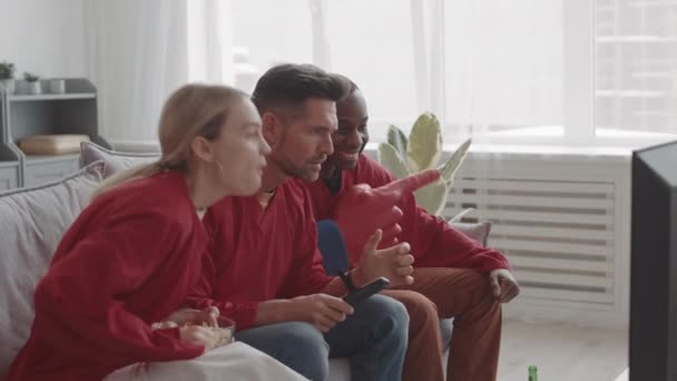 赤い長袖のジャージを着た3人の多様なスポーツファンの中長期的な側面図 自宅でソファに座って 手を上げて お気に入りのチームがゲームに勝つために幸せ — ストック動画