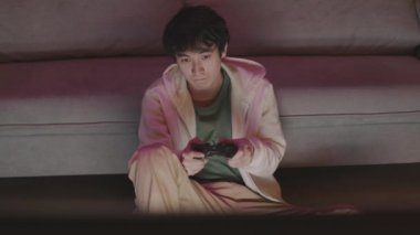 Orta uzunlukta genç Asyalı bir adam yerde oturuyor, kanepeye yaslanıyor, konsol kumandasını tutuyor, geceleri video oyunu oynuyor.