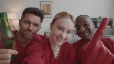 Göğüs hizasında, erkek ve Afrikalı arkadaşlarıyla aynı kırmızı uzun kollu formaları giyen, evinde selfie videosu çeken, sarı saçlı mutlu bir kadın.