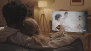 Tanımlanamayan sarışın ve koyu renk saçlı insanların arka koltukta oturup televizyon izlemesi uzaktan kumandayla televizyonun sesini açması.