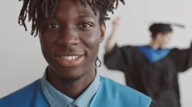 Beyaz arka planda kep ve cüppeli kız arkadaşı neşeyle dans ederken Afro-Amerikan öğrencisinin yakın plan portresi kameraya gülümsüyor.