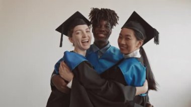 Beyaz arka planda birbirlerine sarılarak kameraya gülümseyen üç beyaz, Asyalı ve Afro-Amerikan mezun öğrencinin orta yavaş portresi.