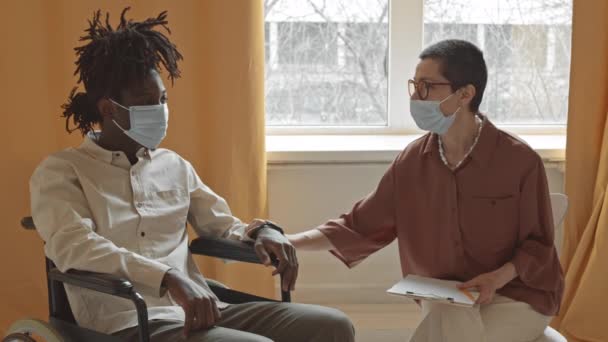 悲しいアフリカ系アメリカ人男性の顔のマスクと車椅子の中での中低速応答でうなずいてセッション中に女性心理学者に耳を傾け — ストック動画