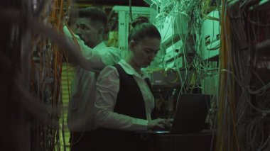 Beyaz erkek mühendis kabloları kontrol ediyor ve genç bayan meslektaşım dar teknik odada taşınabilir bilgisayarda daktilo kullanıyor.