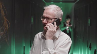 Beyaz gömlek ve gözlüklü, yeşil ışıklı sunucu odasında cep telefonuyla konuşan üst düzey beyaz mühendis.
