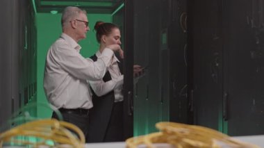 Orta yaşlı, beyaz bir adam ve genç bir kadın resmi giysiler giyiyor, yeşil ışıklı sunucu odasında tartışıyorlar.