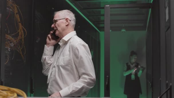 白いシャツとメガネを身に着けているシニア白人エンジニアのウエストアップ 緑の照明サーバールームで携帯電話で話して — ストック動画