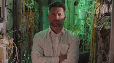 Beyaz gömlek giyen beyaz tenli erkek teknisyen, veri merkezindeki karanlık santral odasında poz veriyor, ellerini göğsüne koyuyor, kameraya bakıyor ve gülümsüyor.