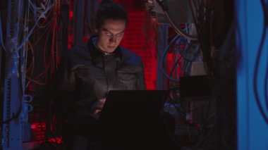 Beyaz tenli gözlüklü genç bir erkek programcı, karanlık santral odasında oturuyor, taşınabilir bilgisayarda kodlanıyor, arka planda iş arkadaşları yürüyor.