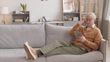 Slowmo of senior man in casual clothes and eyeglasses using digital tablet while relaxing on sofa in bright cozy apartment