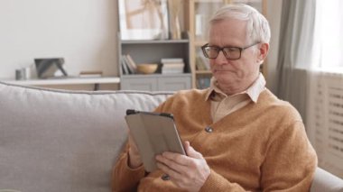 Medium slowmo of cheerful senior man in eyeglasses looking at digital tablet reading news while relaxing on comfortable couch in modern living room