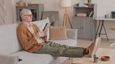 Slowmo of contemporary senior man in casual clothes and eyeglasses talking through video call using digital tablet while sitting on sofa with his legs on coffee table at home