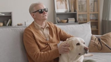 Medium slowmo of blind old man in black eyeglasses petting his Labrador retriever dog while relaxing on sofa in cozy living room