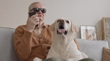 Low-angle medium shot with slowmo of blind old man in black eyeglasses talking through speakerphone on smartphone while petting his cute Labrador retriever dog relaxing at home