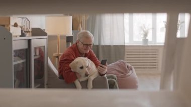 Medium slowmo of cheerful senior man and his cute Labrador retriever video calling on smartphone while relaxing at home together