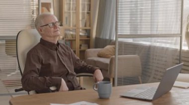 Medium slowmo of cheerful senior office worker or businessman swinging on chair at workplace in modern office
