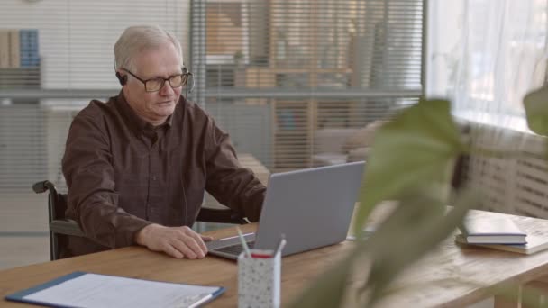 坐在现代办公室轮椅上 穿着时髦便服 头戴耳机 坐在笔记本电脑上的严肃老年商人的慢动作 — 图库视频影像