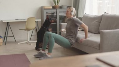 Kafkasyalı genç bir kadının kulaklık, tayt ve tişört giyip evdeki koltuğu kullanarak vücut ağırlık bankı dalışı yaptığı, siyah bir Doberman köpeğinin