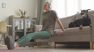 Kafkasyalı genç bir kadının kulaklık, tayt ve tişört giyip evdeki kanepede vücut ağırlık dalışı yaptığı, siyah bir Doberman köpeğinin tenis topu çiğnediği bir sahne.